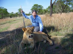 Photo of James Cochran on a lion walk in South Africa, kneeling next to a calm lion.