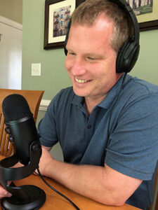 Richard Zink, a white man wearing headphones and a blue shirt, smiles and looks at a microphone. 