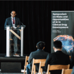 A brown-skinned man stands behind a lecturn and next to a sign that reads Symposium on Risks and Opportunities of Artificial Intelligence (AI) in Clinical Drug Development. The audience is visible, seated in wooden chairs in the front of the image.