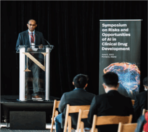 A brown-skinned man stands behind a lectern and next to a sign that reads Symposium on Risks and Opportunities of Artificial Intelligence (AI) in Clinical Drug Development. The audience is visible, seated in wooden chairs in the front of the image.