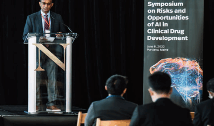 A brown-skinned man stands behind a lecturn and next to a sign that reads Symposium on Risks and Opportunities of Artificial Intelligence (AI) in Clinical Drug Development. The audience is visible, seated in wooden chairs in the front of the image.
