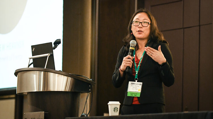 An Asian woman with long wavy hair and black-rimmed glasses holds a microphone and speaks in a conference room.