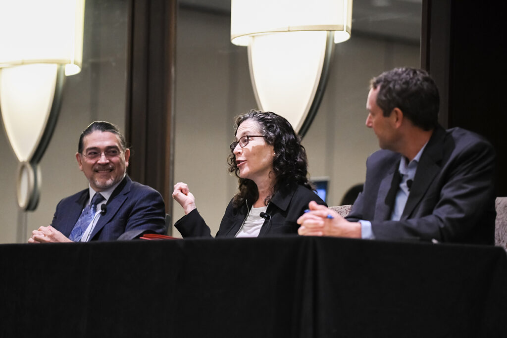 Two men and a woman sit at a table in a conference room. The woman is in the center and wears glasses and has long curly dark hair. To her left is a Latino man wearing glasses and a suit and tie. The man to her right is out of focus and looking at the woman.