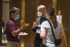 Three white people hold plates of food and cups and chat while wearing medical-grade face masks. Two men are in the rear of the photo, and a woman seen from the back is in the foreground. She is wearing a ponytail and a fanny pack as a cross-body bag.