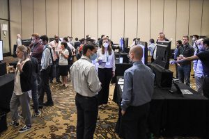 A room full of people, some wearing face masks and some not, some eating and drinking, in a hotel conference room with several rows of TVs showing people's research posters.