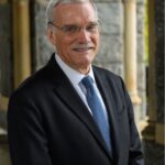 A white man with gray hair, a gray mustache, and glasses smiles. He is wearing a blue suit and lighter blue tie.