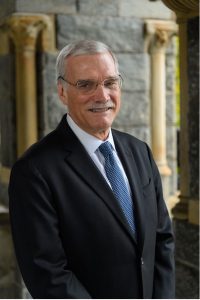 A white man with gray hair, a gray mustache, and glasses smiles. He is wearing a blue suit and lighter blue tie. 