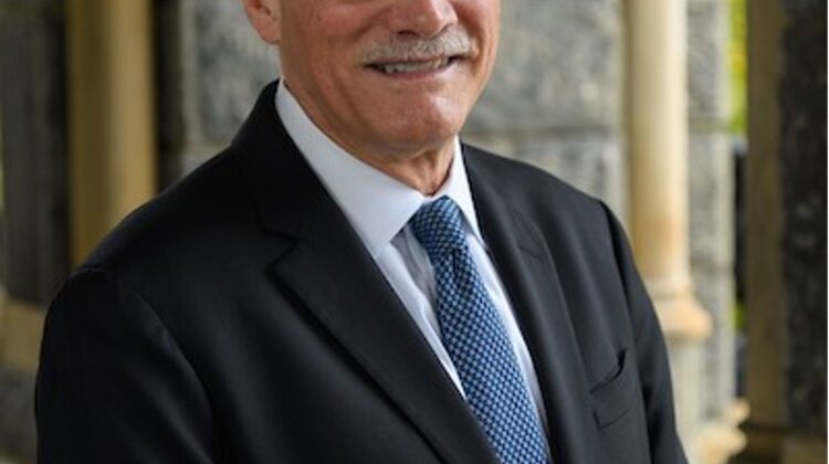 A white man with gray hair, a gray mustache, and glasses smiles. He is wearing a blue suit and lighter blue tie.