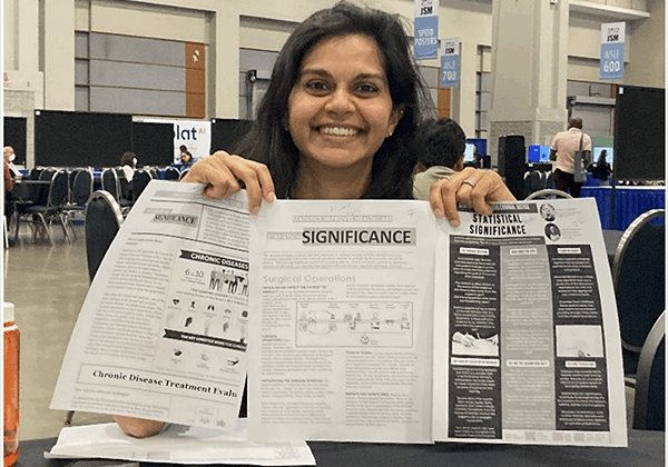 A brown-skinned woman with dark hair smiles as she holds up the winning Statistcal Significance entries.