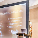 A white woman stands behind a lectern to the right of the frame. To her left is a screen with text reading "Presidential Initiatives.' The categories on the slide are Leadership and Community Analytics.