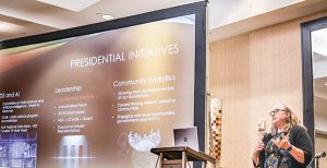 A white woman stands behind a lectern to the right of the frame. To her left is a screen with text reading "Presidential Initiatives.' The categories on the slide are Leadership and Community Analytics.