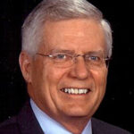 An older white man with frameless glasses smiles. He has gray hair.