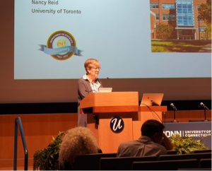 A white woman with short hair stands behind a presentation screen at a lectern. 