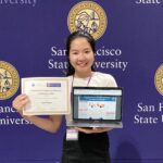 Lana Huynh, an Asian woman, stands in front of a purple backgound that has the San Francisco State University logo repeating across it. She is smiling and holding a laptop displaying her research and a certificate.