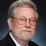 A white man with gray hair and a gray beard wearing wire-rimmed glasses