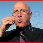 A white, bald man wearing frameless glasses blows bubbles, which surround him. He is wearing a grey-and-black striped tie and a black shirt against a blue sky.