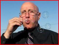 A white, bald man wearing frameless glasses blows bubbles, which surround him. He is wearing a grey-and-black striped tie and a black shirt against a blue sky.  