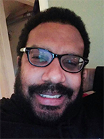 A Black man wearing glasses with dark frames and a mustache and beard and short hair smiles