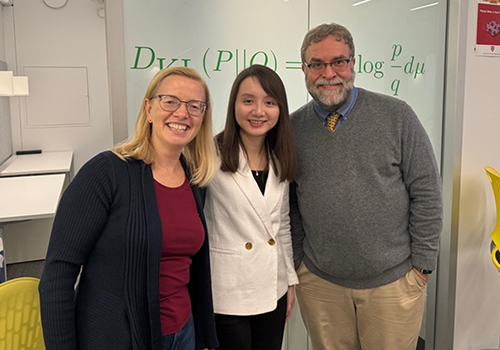 A white woman with long blond hair and glasses stands next to an Asian woman with long dark hair who is next to a white man with a beard and glasses. They are all smiling. 