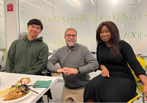 A white man with gray hair and and beard and glasses sits between an an Asian man to his left and a Black woman with long, straight dark hair on his right. 