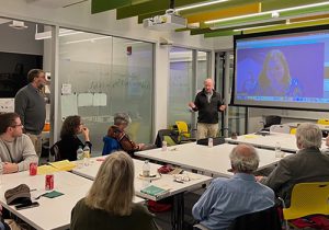 An older white man stands by a projection screen lecturing to a conference room.