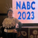 Arinjita standing in front of a sign that reads NABC 2023. She is wearing a headdress and a lace covering over her shoulders.