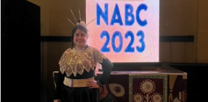 Arinjita standing in front of a sign that reads NABC 2023. She is wearing a headdress and a lace covering over her shoulders.