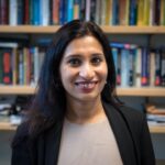 An Indian woman with long dark hair, wearing a black suit jacket and a tan shirt, smiles. 