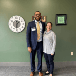 A Black man and white woman stand in front of a green wall. The man wears a dark suit and the woman wears a grey shirt.