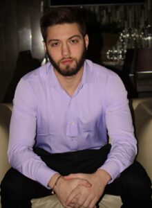 A dark-haired man with a beard wearing a light purple shirt leans forward, hands clasped.