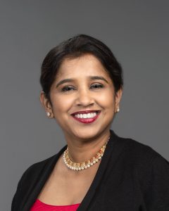 A southeast Asian woman with short dark hair, gold earrings and necklace, and a suit jacket, smiles widely
