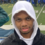 Wearing hoodie, standing in bleachers, football game in background.