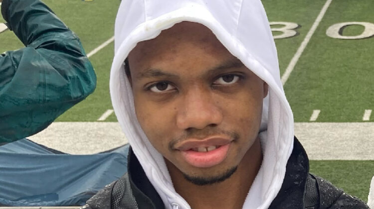 Wearing hoodie, standing in bleachers, football game in background.