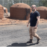 Baiocchi is standing in front of mounds of sand, walking on a dusty road. He is wearing boots, long pants, short sleeve shirt and glasses.