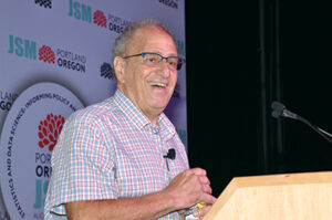 An older man wearing glasses and a plaid shirt stands behind a lectern. He is grinning.