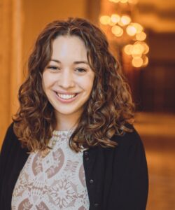 Moira Skelley's headshot. She has shoulder-length curly brown hair and is wearing a black cardigan with a white lace top.