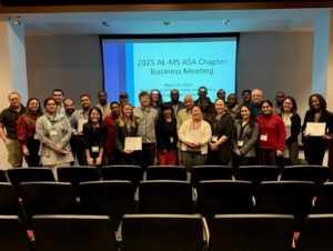 A large group of approximately 30 attendees poses for a group photo at the 2025 AL-MS ASA Chapter Business Meeting in a lecture hall, with several people holding certificates in front of a projection screen displaying the event details.