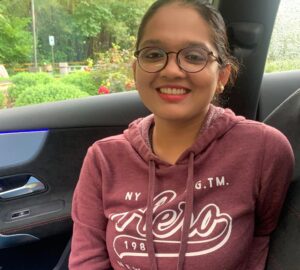A smiling brown-skinned woman with her hair pulled back is wearing glasses, a brick-colored sweatshirt, and glasses.