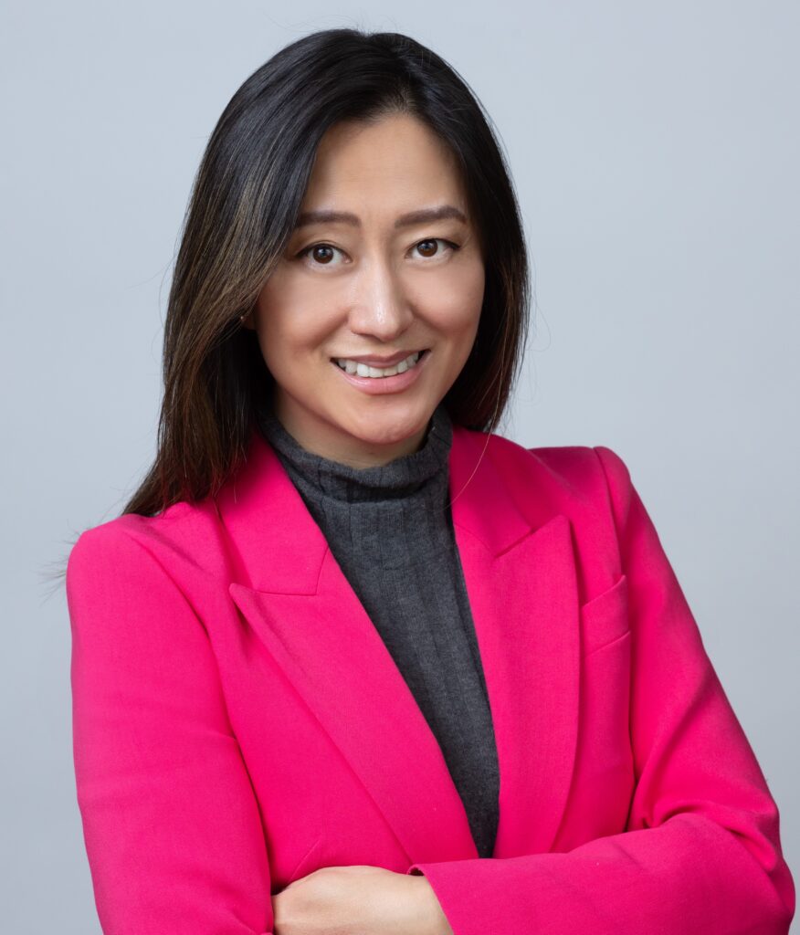 An Asian woman with long dark hair, wearing a hot pink blazer and a gray turtleneck, smiles