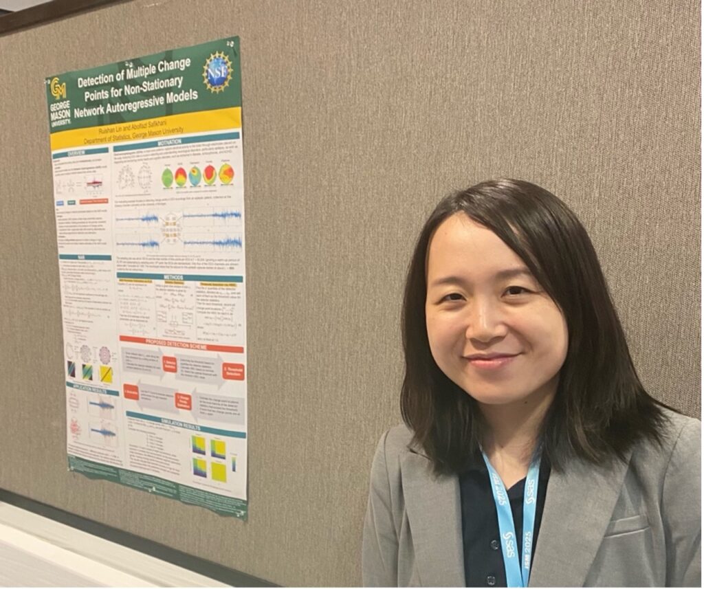 An Asian woman with shoulder-length black hair, wearing a gray blazer and a dark shirt, stands near a poster presentation