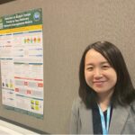 An Asian woman with shoulder-length black hair, wearing a gray blazer and a dark shirt, stands near a poster presentation