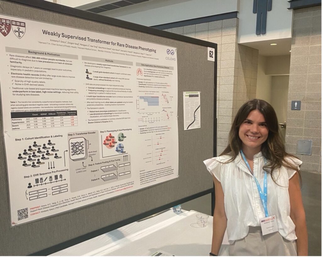 A white woman with mid-length dark hair, wearing a white blouse and khaki pants, stands near a poster presentation