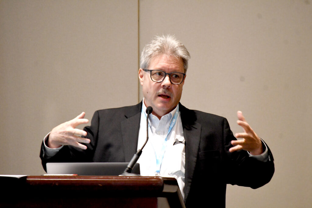 A white man with gray hair behind a podium gestures with wide-open arms