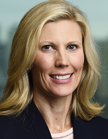 A white woman with blonde hair, wearing a dark suit jacket, smiles