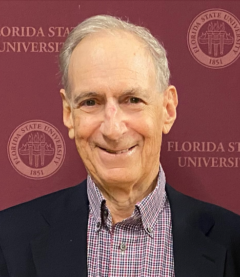A white man in a gingham shirt and jacket with white hair smiles