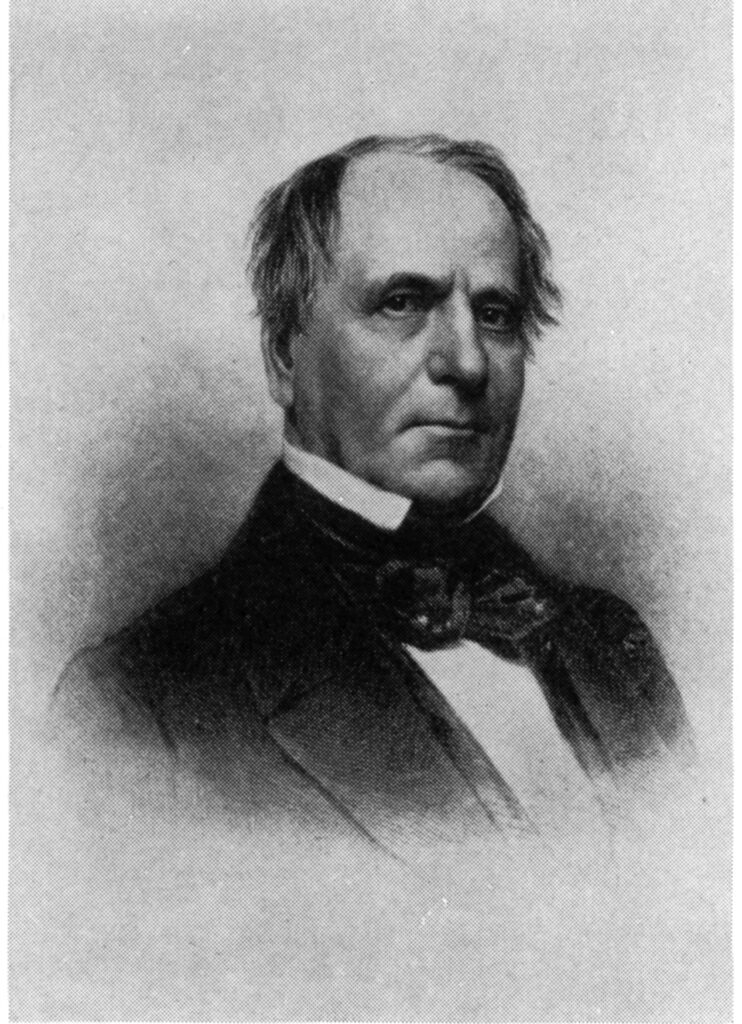 A black and white image of a man with wispy hair, wearing a tie and jacket.