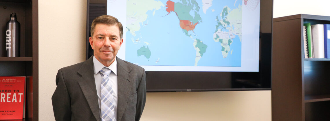 A white man in a gray suite with a gray geometric tie in front of map on a TV with a world map on it.