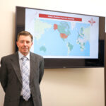 A white man in a gray suite with a gray geometric tie in front of map on a TV with a world map on it.