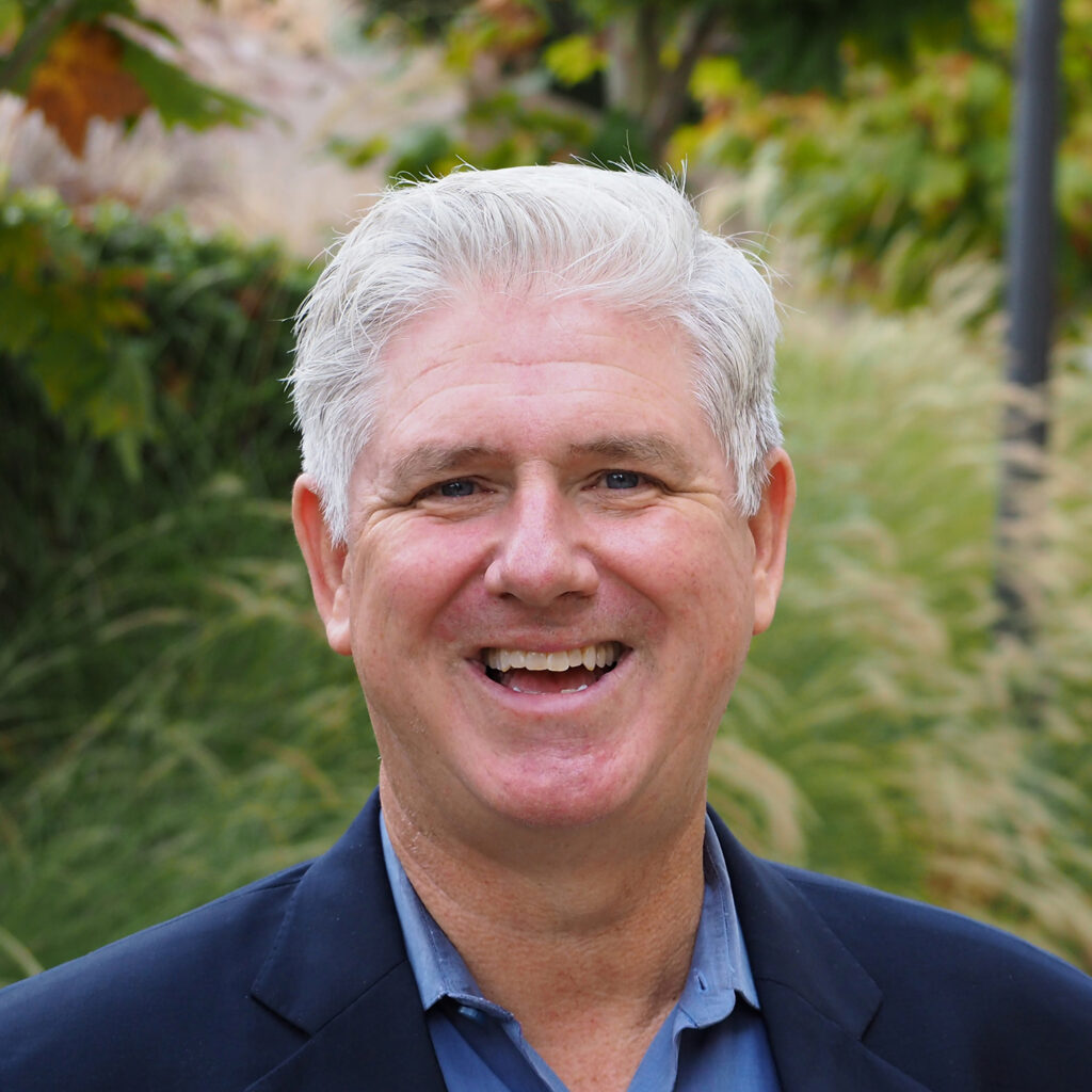 Rob Gould smiles at the camera. He is wearing a blue shirt and jacket and has short white hair.