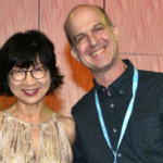 An Asian woman with short dark hair and glasses stands with a white man in a dark patterned shirt with short hair.
